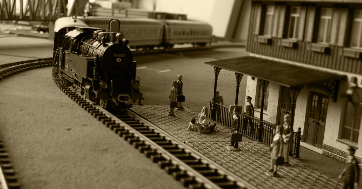 A model locomotive pulls up to a small train station. There are multiple people figurines awaiting its arrival.