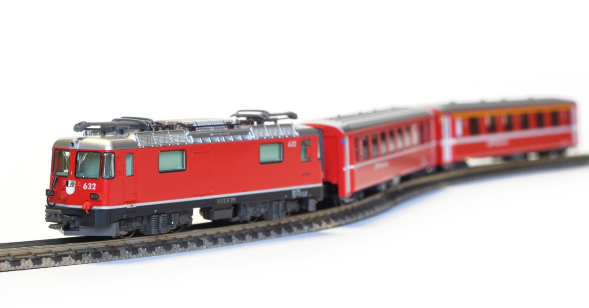 A scale model of a three car, red train on a piece of track. The train is set against an empty white background.