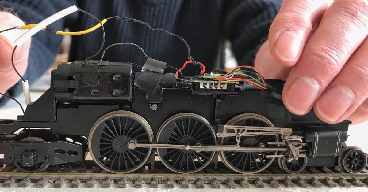 A hobbyist inspecting the wiring inside of a black model train. There are several wires sticking out of the top of the train.