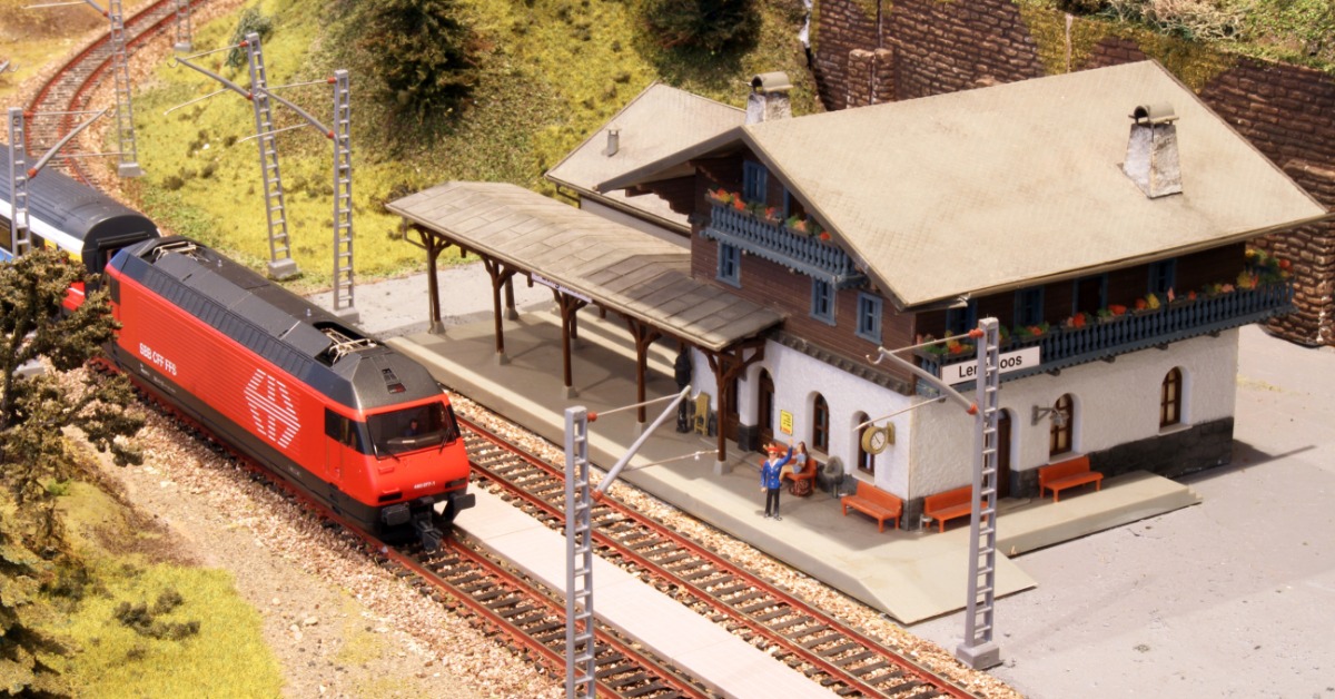An orange model train pulling into a small model train station on the side of the track. Tiny figurines are in the station.