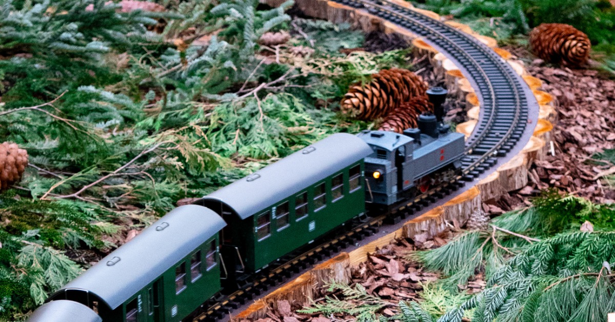 A dark green model train making its way through a model train track that is built into a garden outside.