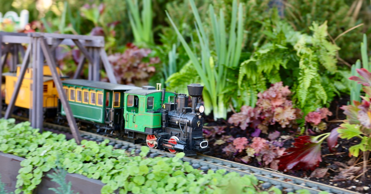A model train with several multicolored train cars going across a bridge that is built over a garden.