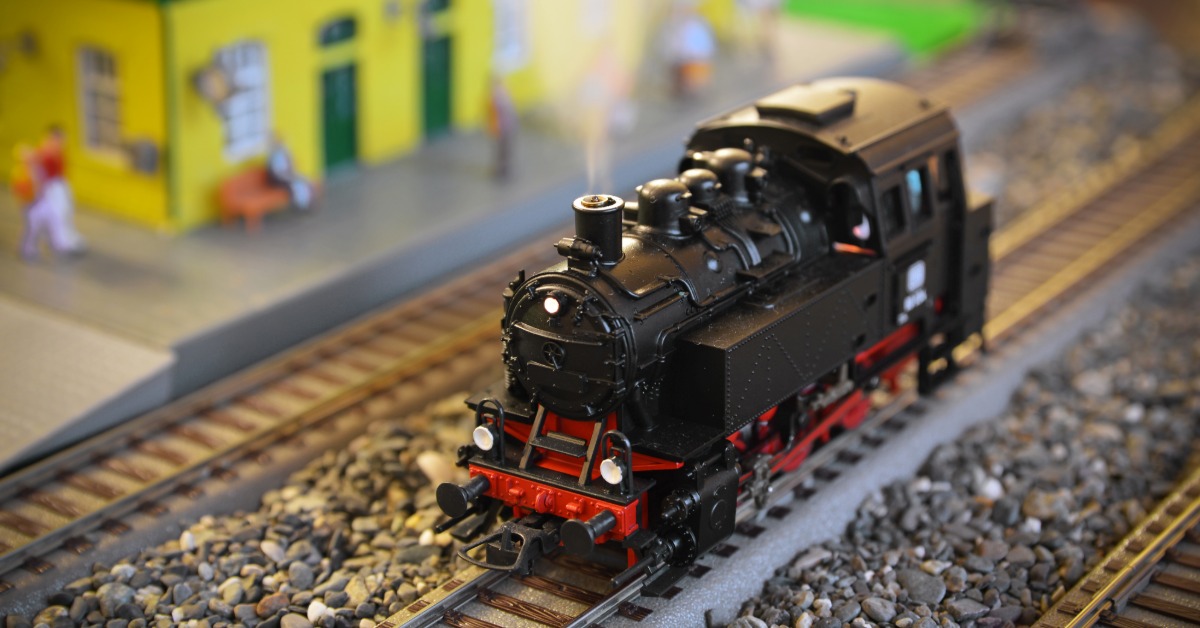 A small model steam locomotive on a track in front of a small train station. The track is on top of tiny pebbles.
