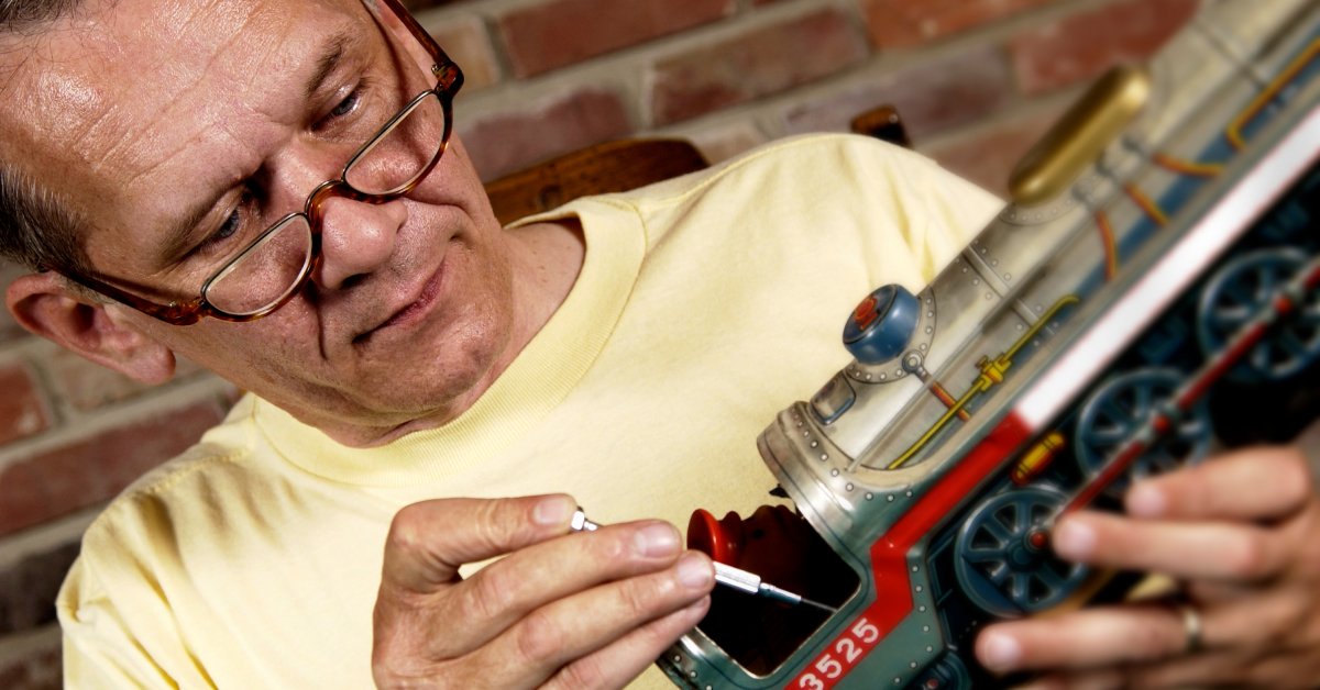 A person holds a model train in their left hand and uses a tool in their right hand to work on the train car.