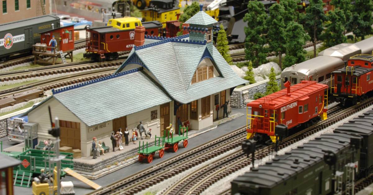 A model train station has multiple train cars on the tracks and small models in the background of trees and people.