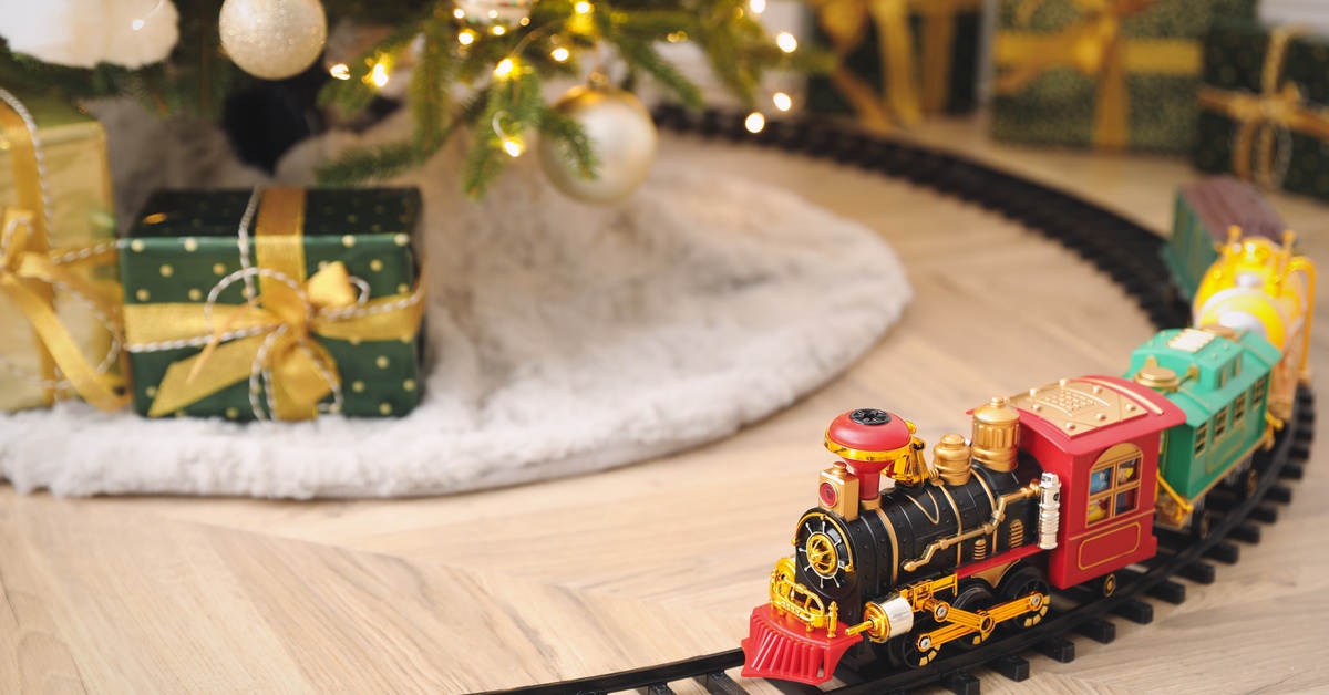 A small toy trainset is going around a Christmas tree. The tree is lit and decorated, and the train is colorful.