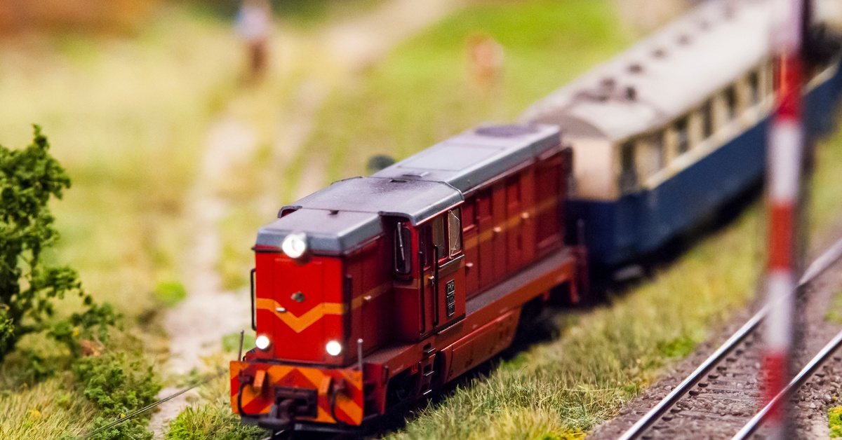 A close-up of a model train driving down a small railroad track. There is some blurry grass and figures behind the train.