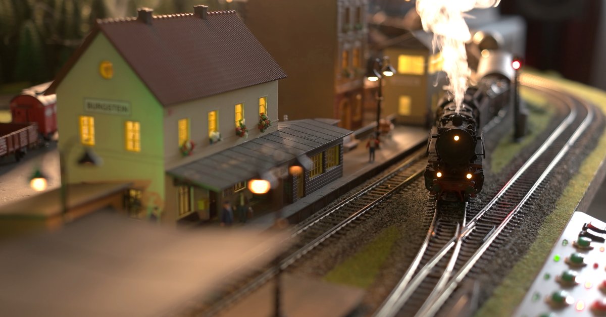 A small model train blows steam out of the front engine while it departs from a small model train station.