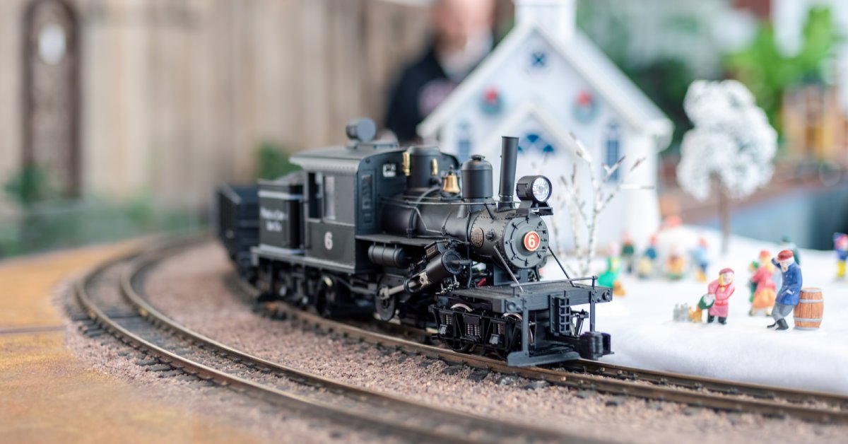 A small model Christmas train driving on a train track next to a small model field of snow and people.
