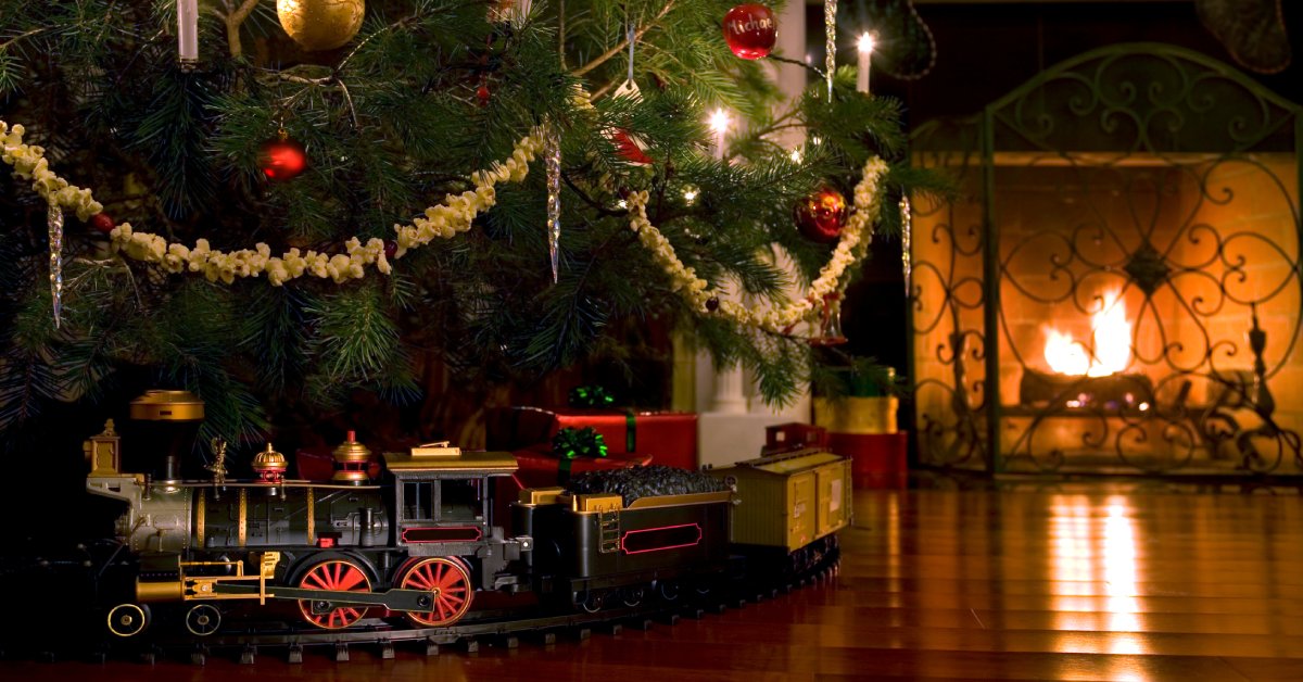 A Christmas tree with a small model train circling the base. There's a large fireplace near it.