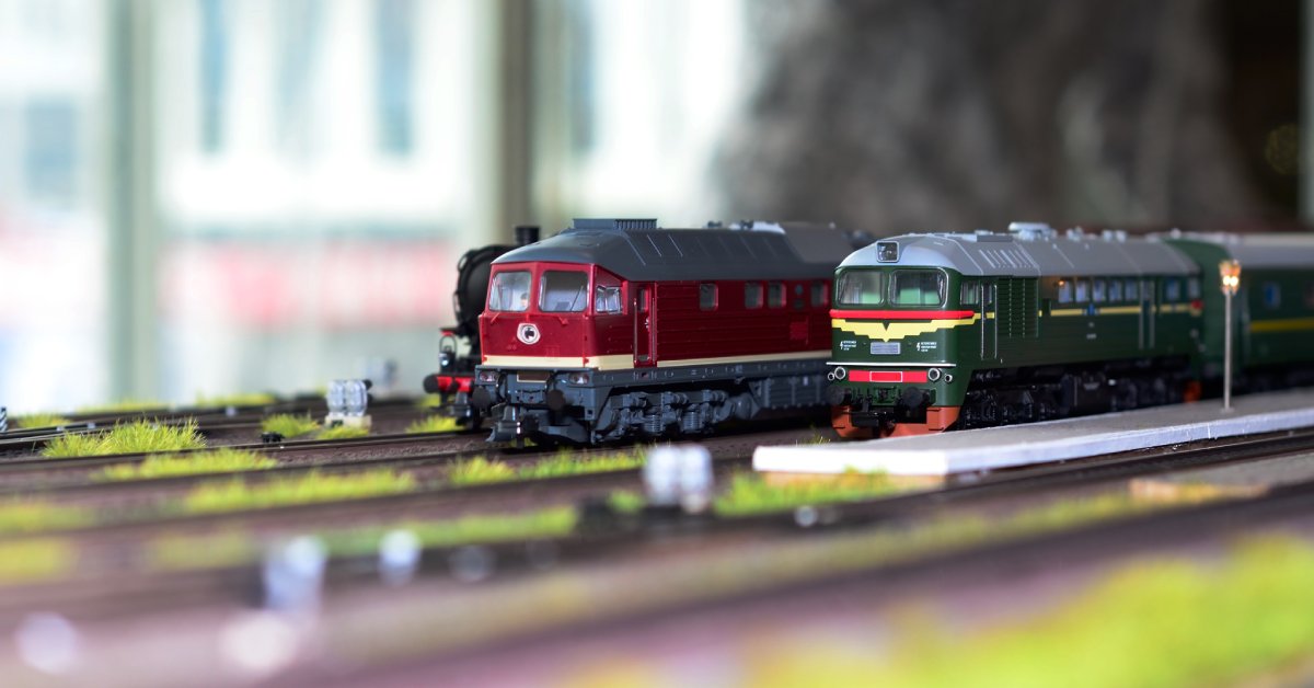 A few small model trains driving through a tunnel and down a railway with grass in between the tracks.