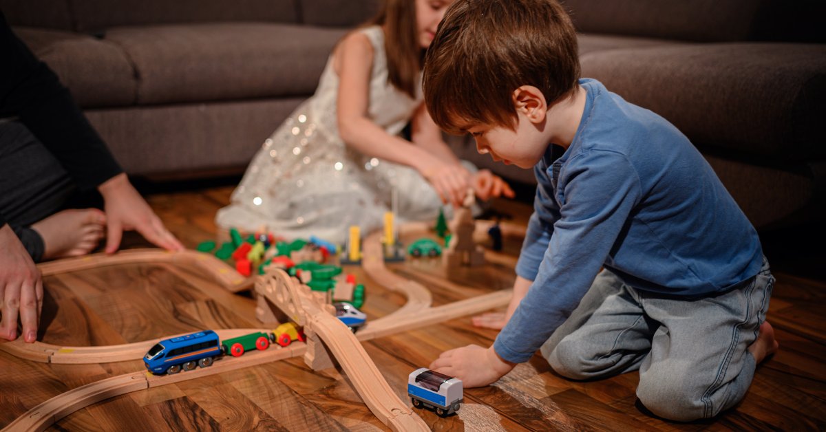 A parent sits on the ground with two children while they all play with small wooden toy trains together.