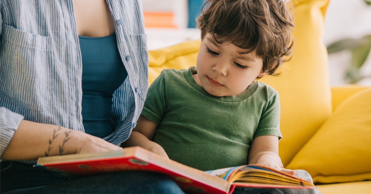 A parent and their child sit together on a couch and they read a small book with a red cover together.