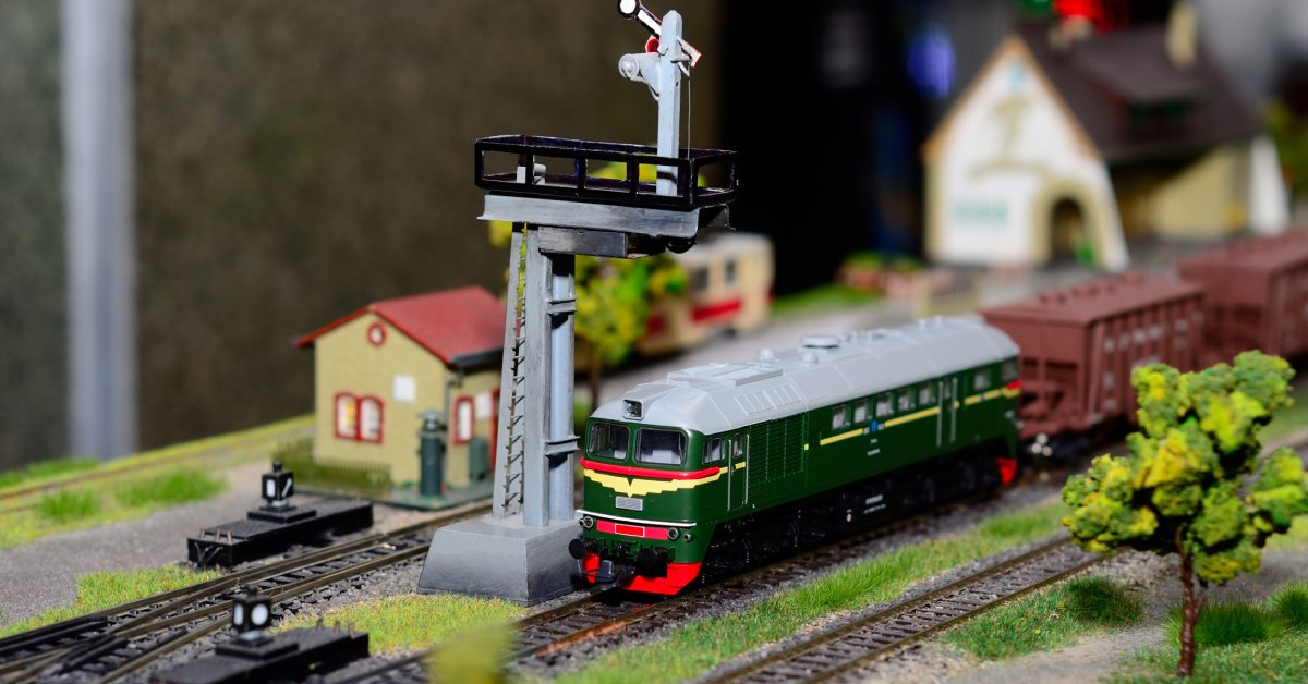 A close-up of a green model passenger train car on the track, driving past the railway rail guard arm.