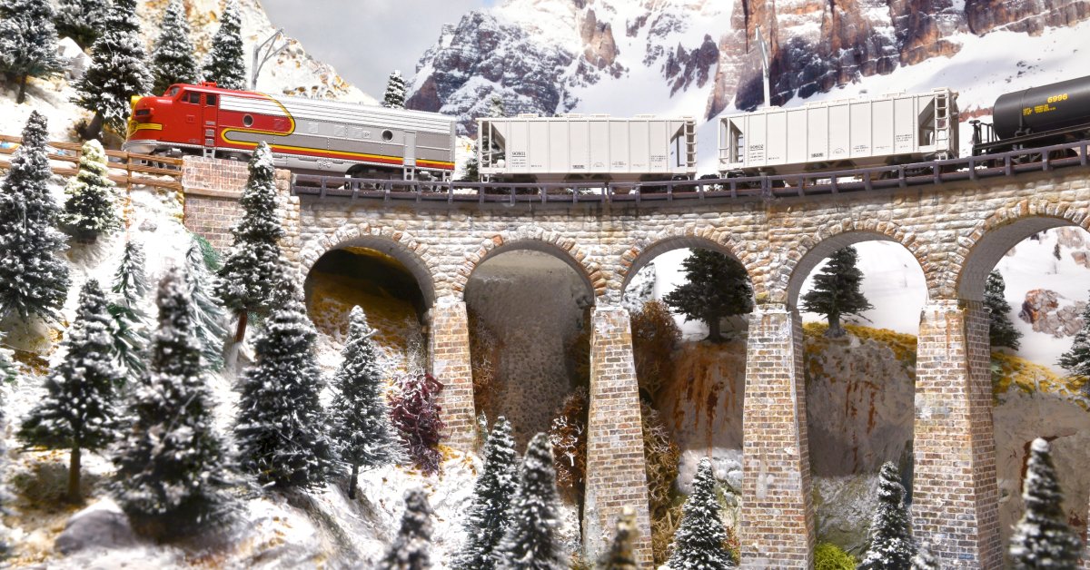 A miniature model train drives over a bridge, navigating a snowy landscape with lots of mountains and trees.