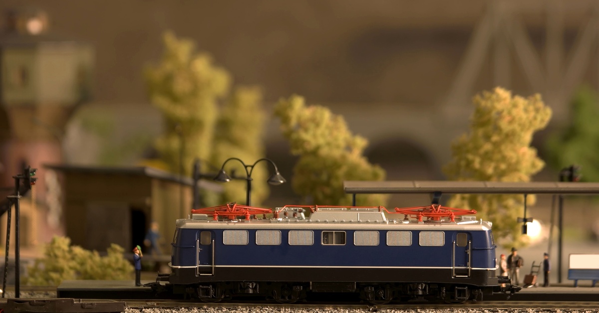 A blue-and-red model train sits on a railroad track in front of miniature people, trees, and lights.