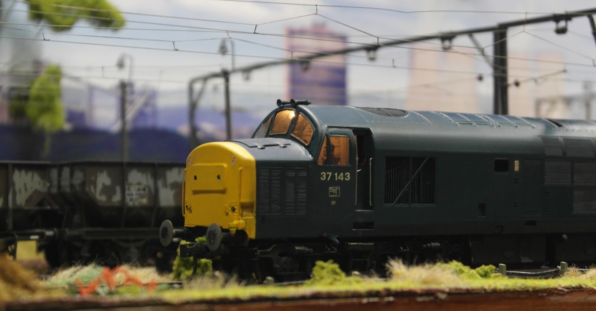 A class 37 model train drives down a model train track. The train is green and yellow and there is other scenery behind it.