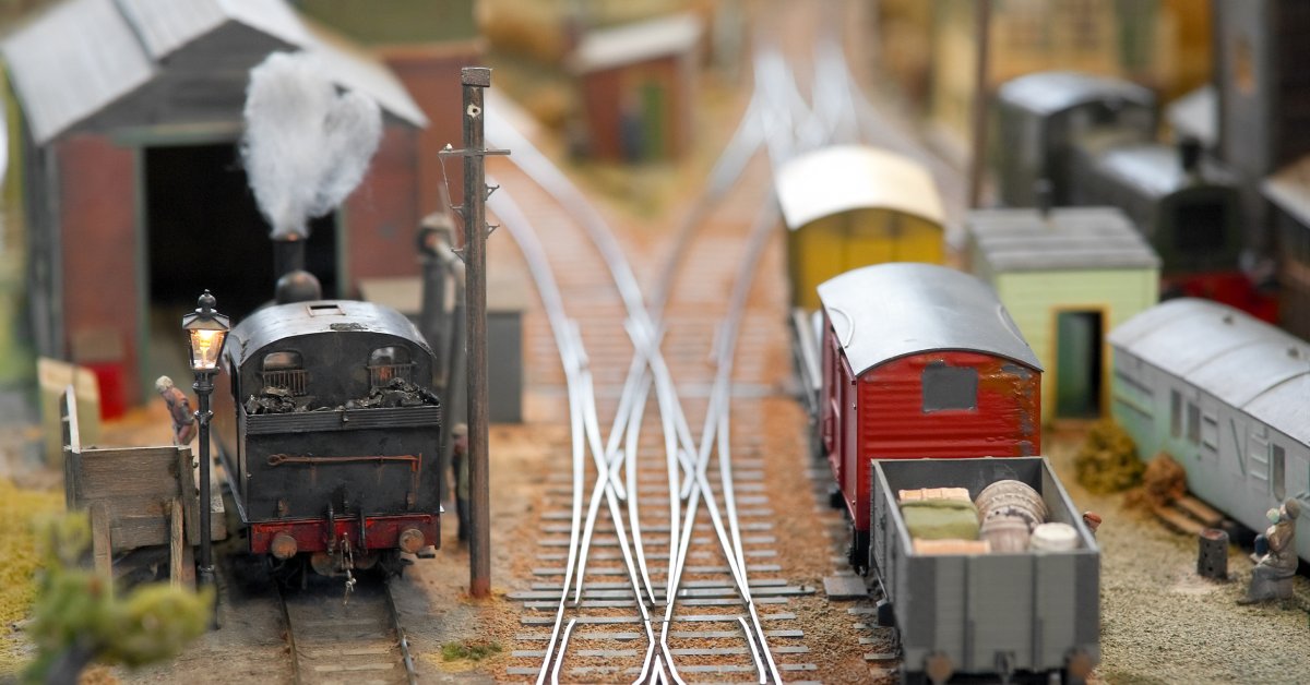 A close up of two model trains sitting on an interconnected railroad track in the middle of a model freight yard.
