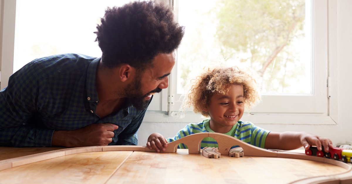A father and a son sit at a table together and the kid plays with a small toy train set, moving it over a wooden track.