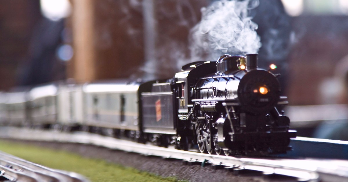 A small model steam train with multiple cabins traveling down a model railroad track, puffing up steam behind it.
