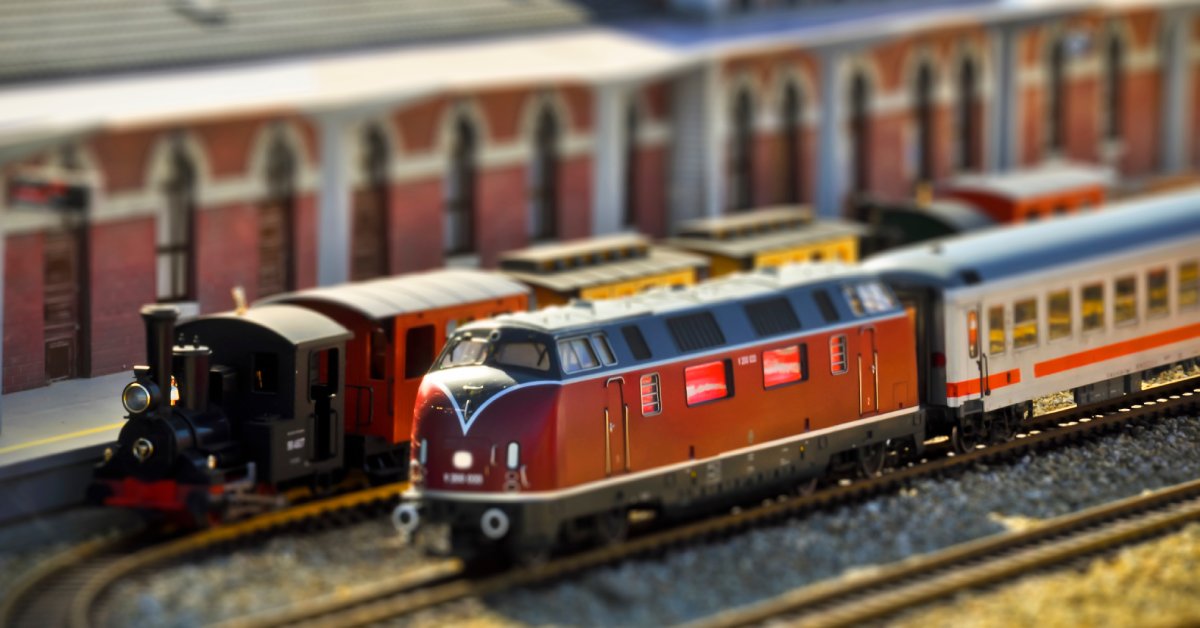 A close-up of a model railway system with a steam car and passenger cars linked on the track, driving past buildings.